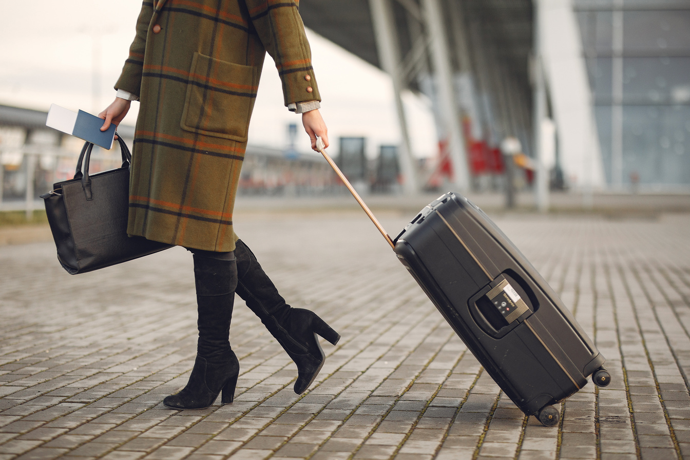 Stylish woman with suitcase and bag walking on street near modern airport terminal